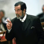 Man in a dark suit and white shirt speaking passionately, gesturing with a pen at a podium in a formal setting