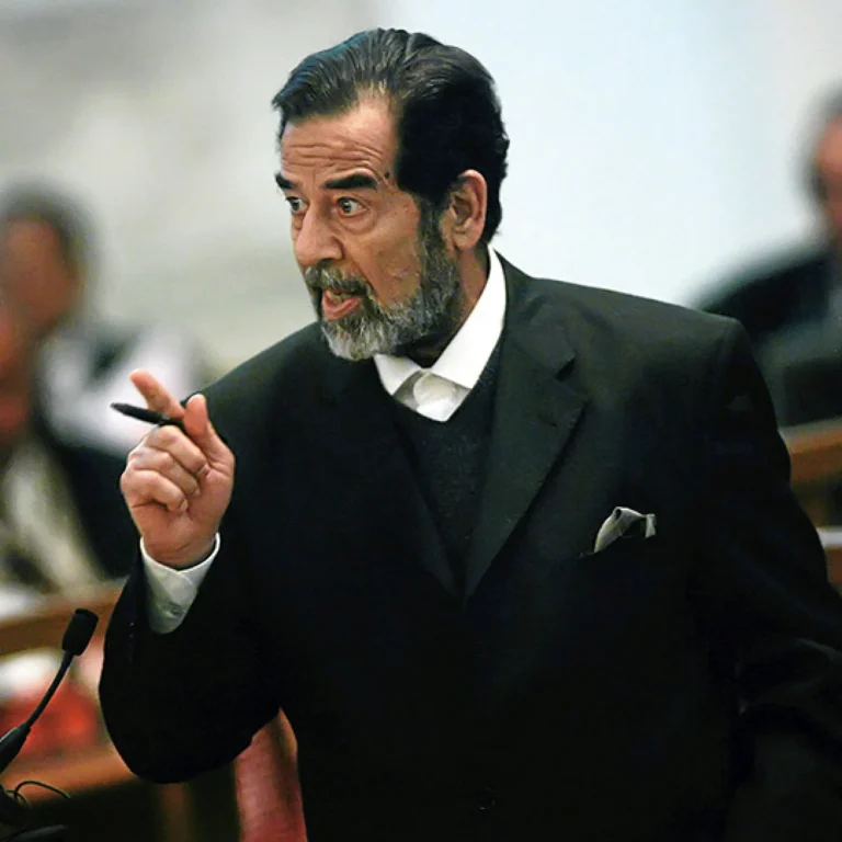 Man in a dark suit and white shirt speaking passionately, gesturing with a pen at a podium in a formal setting
